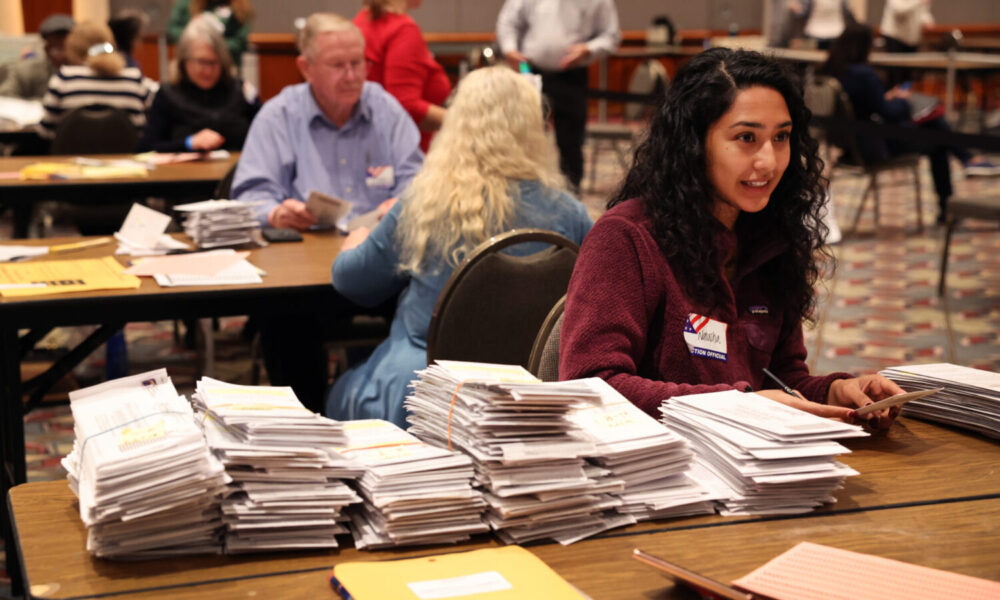 A woman sitting at a tabl;e covered in ballots. Other tables with ballots and people are visible in the background.