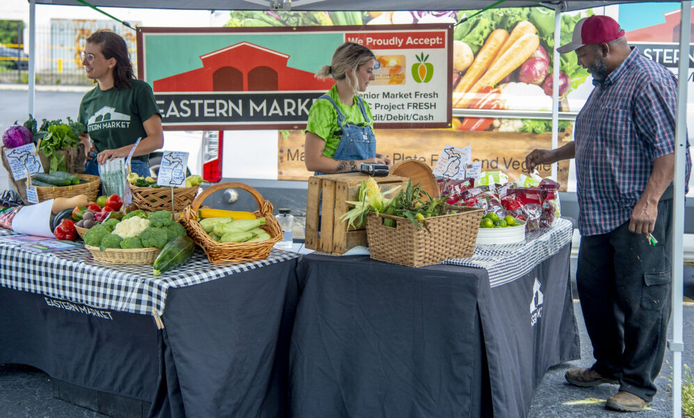 photo of two women selling fruits and vegetables from a stand under a tent at a farmers market, while a male customer looks over the merchandise