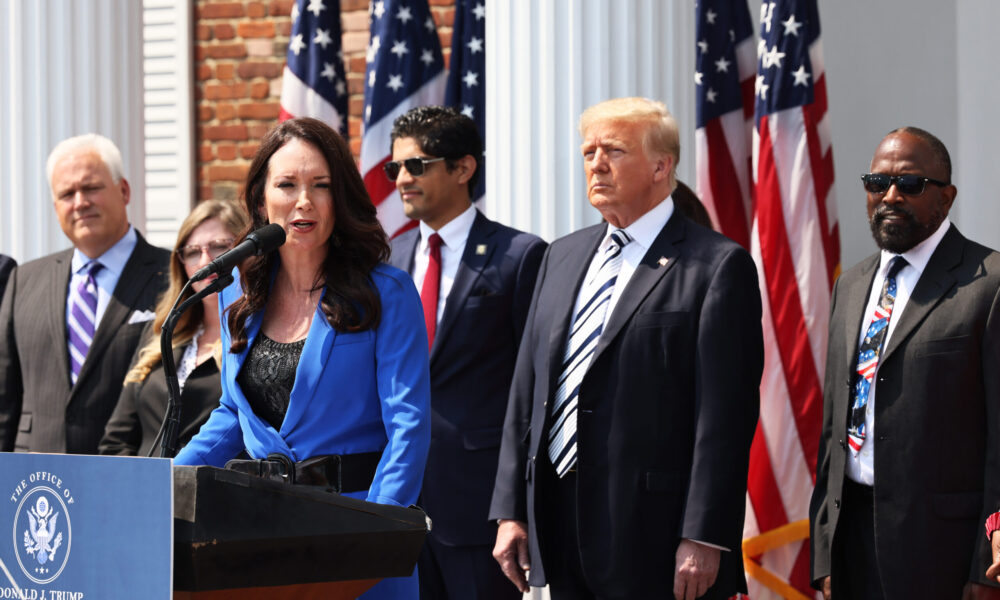 Brooke Rollins stands at a podium with a presidential seal on it and speaks to an unseen audience through a microphone; behind her are a group of people including Donald Trump, and a set of four US flags