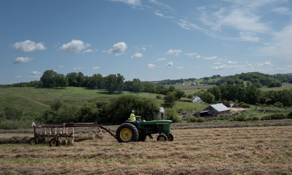 photo of a farmer on a tractor pulling a hay rake through a field; in the background are a farmhouse, low green hills, and blue sky