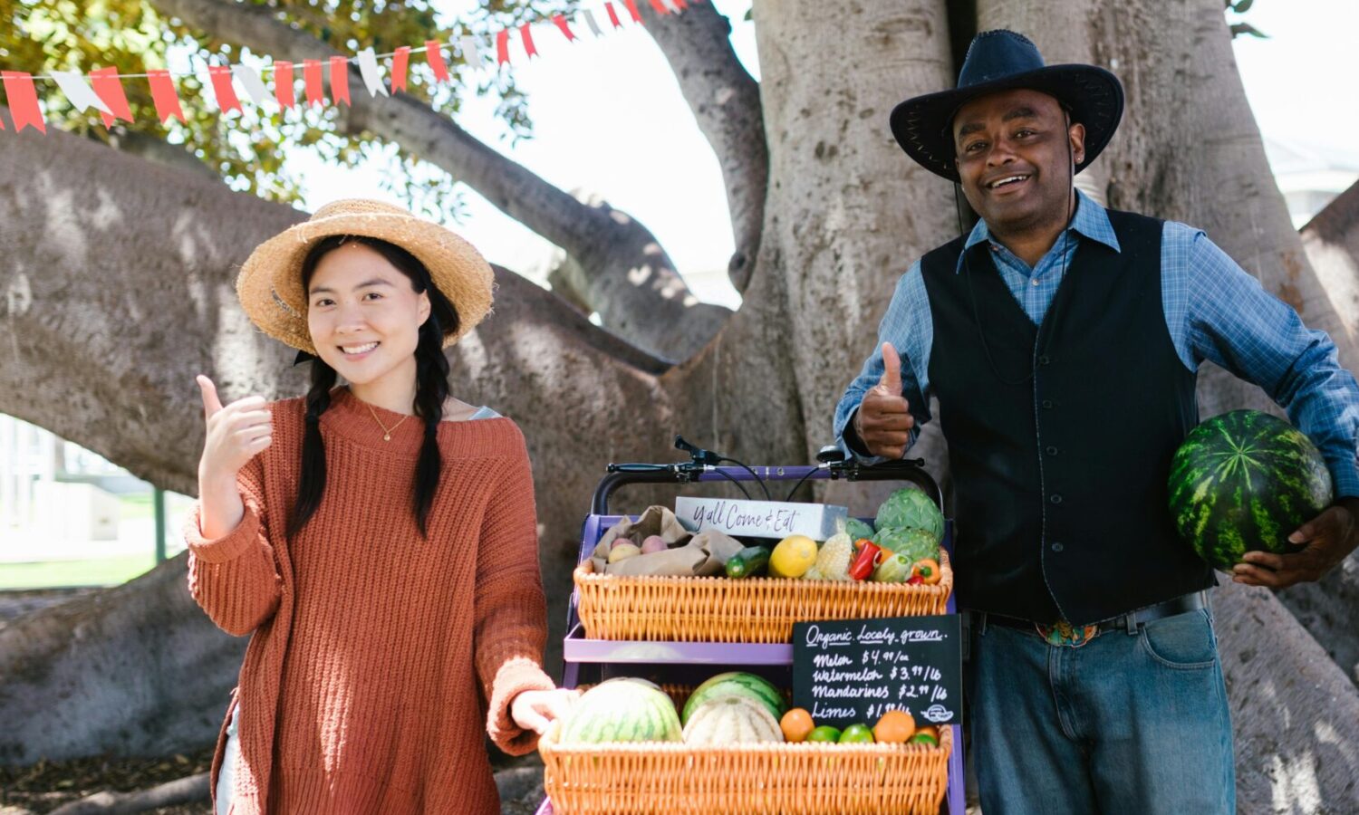 photo of a woman and man standing on either side of a cart with multiple baskets of vegetables for sale, the woman has pigtails and a sunhat while the man is wearing a black cowboy hat and black vest, and both are giving a thumbs-up and smiling at the camera