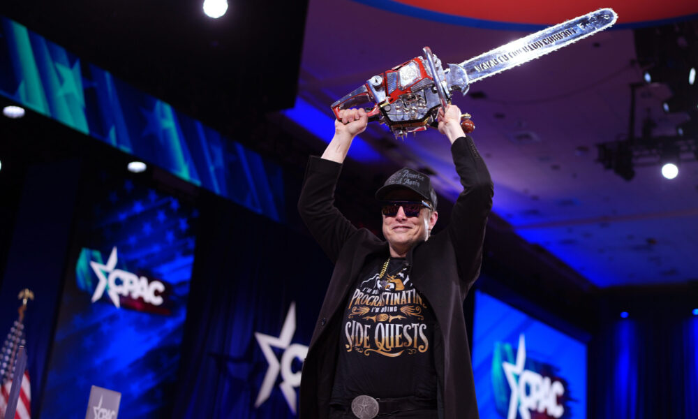 photo of Elon Musk on stage at a rally of the Conservative Political Action Committee. Dressed in all black and wearing sunglasses, Musk smiles as he holds a chainsaw over his head with both hands