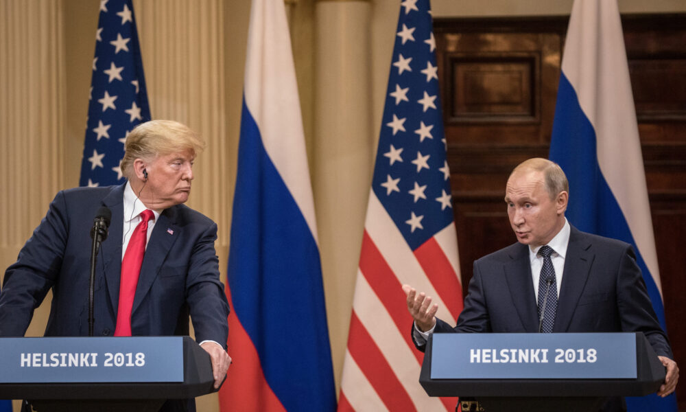 photo of Russian President Vladimir Putin and US President Donald Trump at a press conference during their 2018 summit; they stand at separate podiums bearing the label "Helsinki 2018" and with Russian and US flags behind them. Putin is speaking and Trump is listening to him