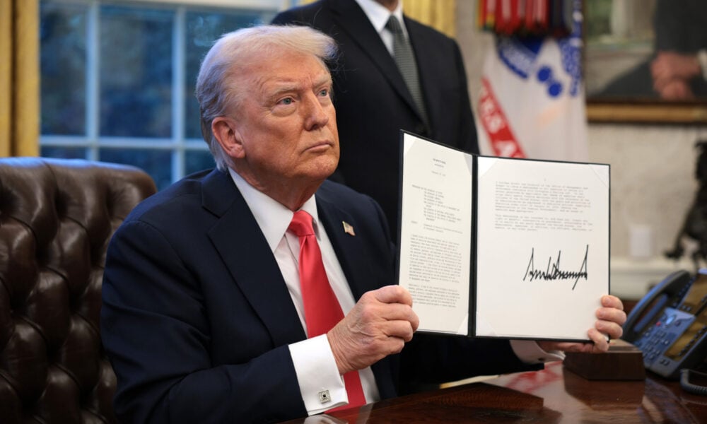 President Trump, in the Oval Office, holds up a signed executive order