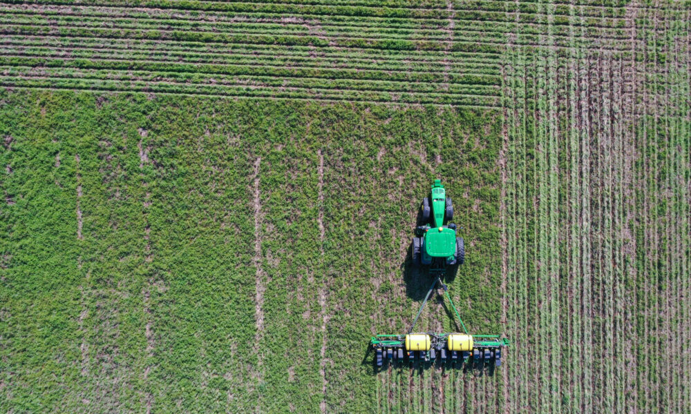 aerial photo looking down on a green tractor pulling fertilization equipment through a cornfield. The tractor's path has left a pattern of intersecting lines in the dirt.